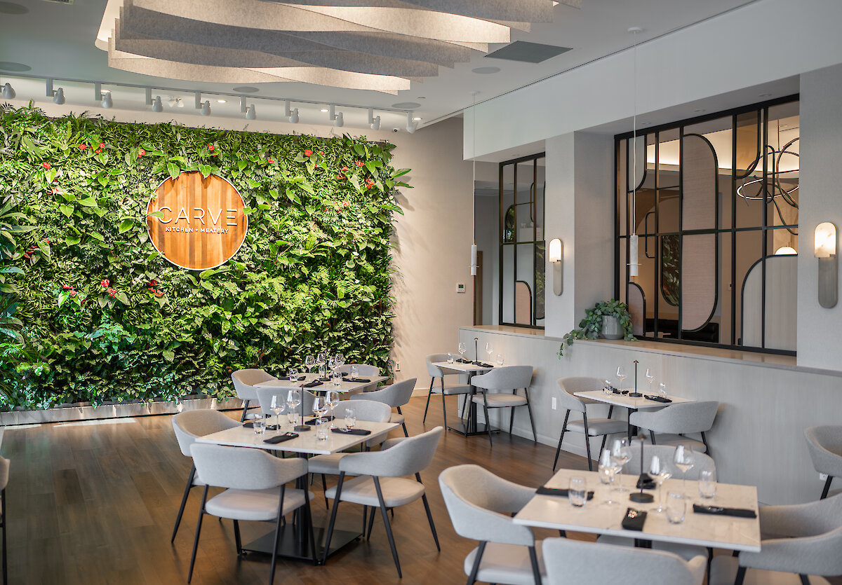 A glimpse inside the neutral coloured restaurant with tables for 4 with modern, c-shaped dining chairs, and a living wall.