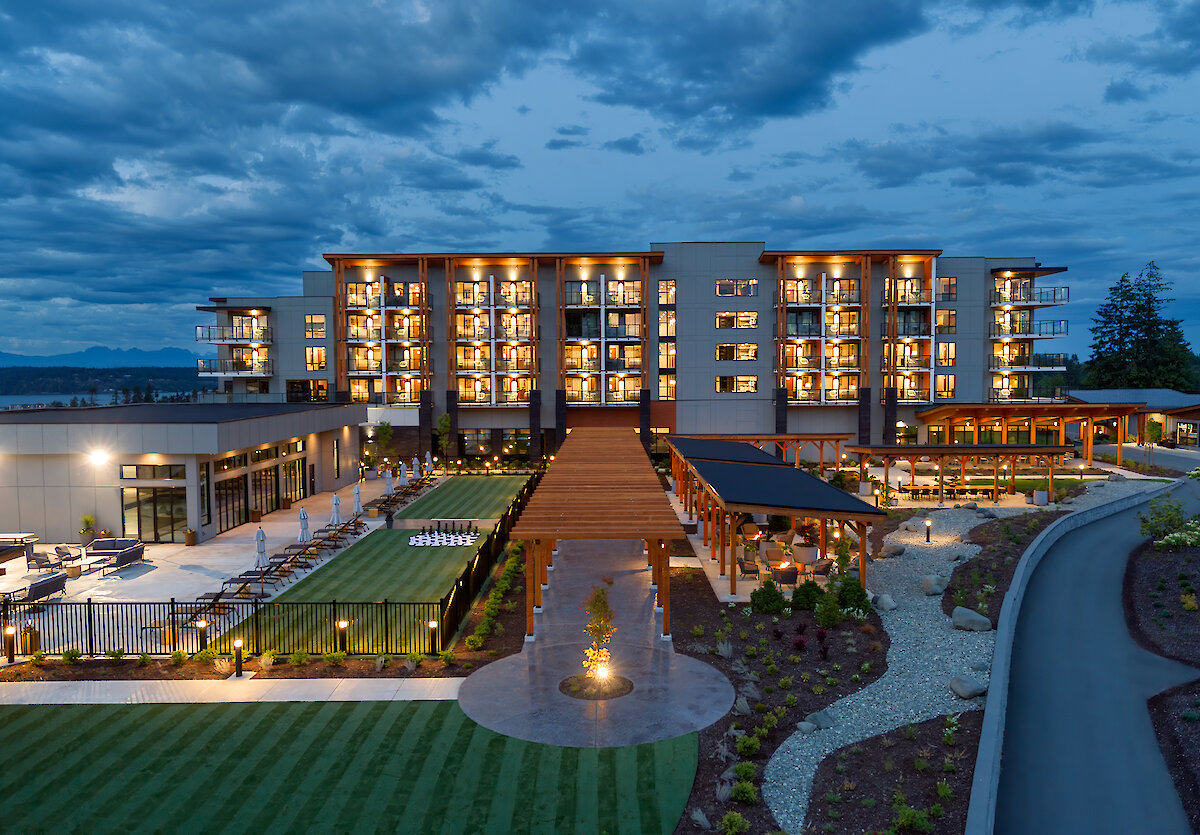 Back side of Naturally Pacific Resort with darkened cloudy skies, rooms lit up, striping on the lawn, yard games and cart paths with distant mountains and ocean in the background