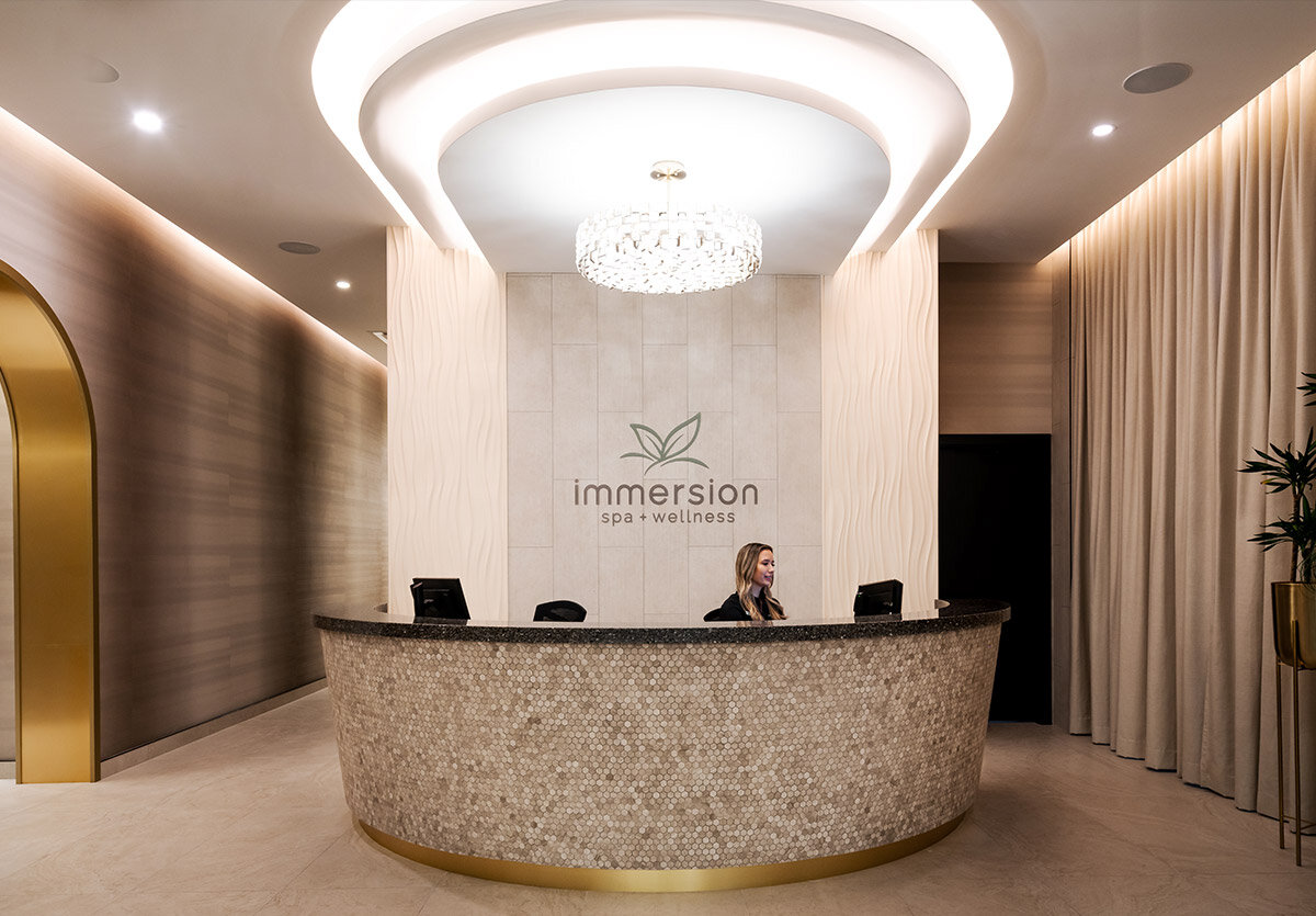 luxurious entrance of immersion spa and wellness looking at the tiled front desk with the logo on the wall and a receptionist at the desk sitting at her computer